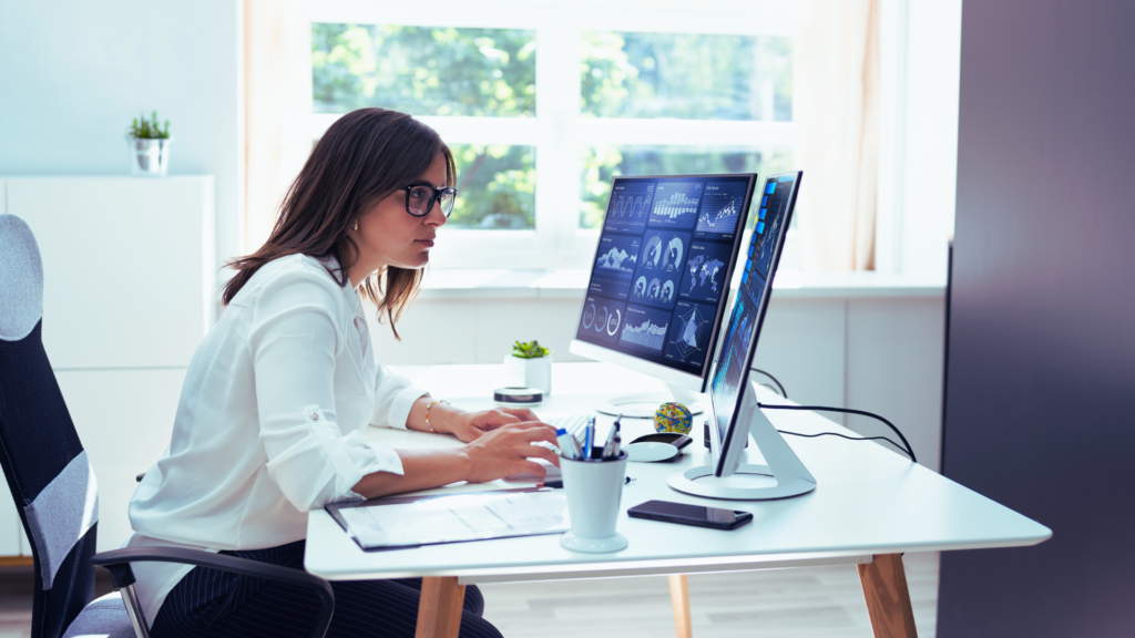 Woman in glasses and a white shirt sitting in an office chair using google analytics for interior design websites on 2 computer monitors on a modern desk.